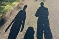 Drie schaduwen vallen over een zonnig asfaltpad, met groen langs de linkerrand.