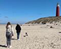 Twee mensen lopen over een breed zandstrand richting een duin met een hoge rode vuurtoren.