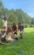 Vijf mensen zitten of hurken op een groen grasveld rond een zwart-witte hond met een blauwe lijn.